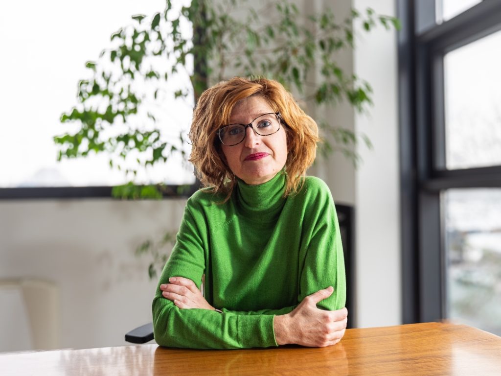 Isinet Consulting Professionista seduto a un tavolo in un ambiente luminoso, indossa un maglione verde e occhiali, con piante sullo sfondo.