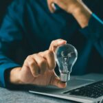 Isinet Consulting Un professionista in camicia blu tiene in mano una lampadina accesa vicino a un computer portatile, simbolo di innovazione e ricerca tecnologica.