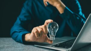 Isinet Consulting Un professionista in camicia blu tiene in mano una lampadina accesa vicino a un computer portatile, simbolo di innovazione e ricerca tecnologica.
