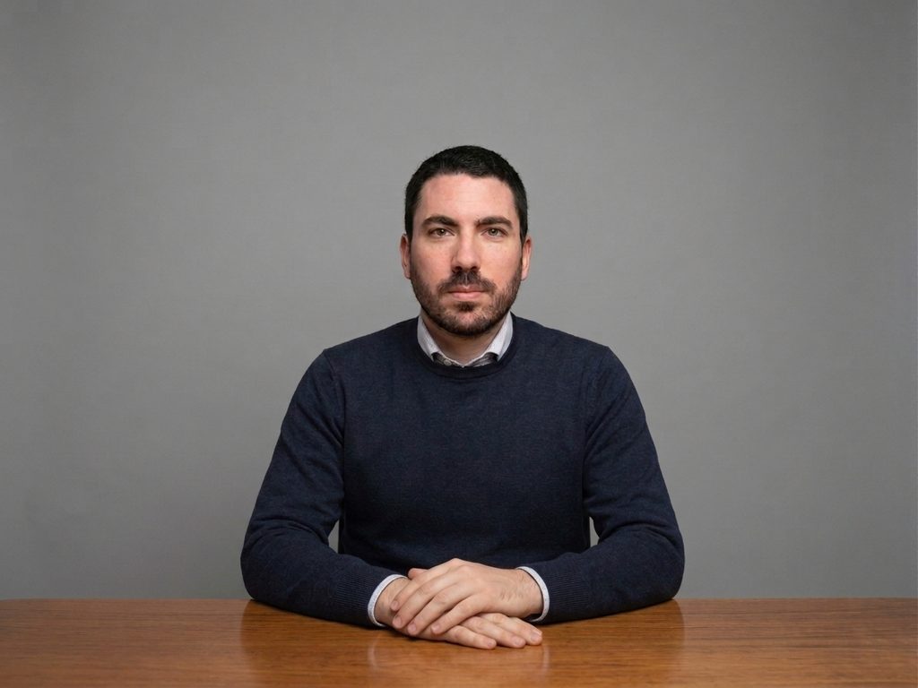 Isinet Consulting Un uomo con barba e maglione blu seduto a un tavolo di legno, ritratto professionale per L'Ufficio Studi su sfondo grigio neutro.